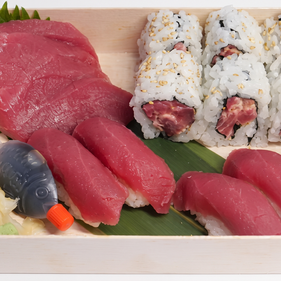 Wooden tray of tuna sushi, sashimi, sesame-topped rolls, pickled ginger, wasabi, and a soy sauce bottle.